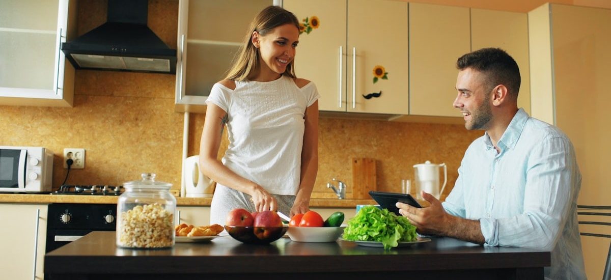 Attractive couple chatting in the kitchen early morning. Handsome man using tablet while his girlfriend cooking breakfast at home